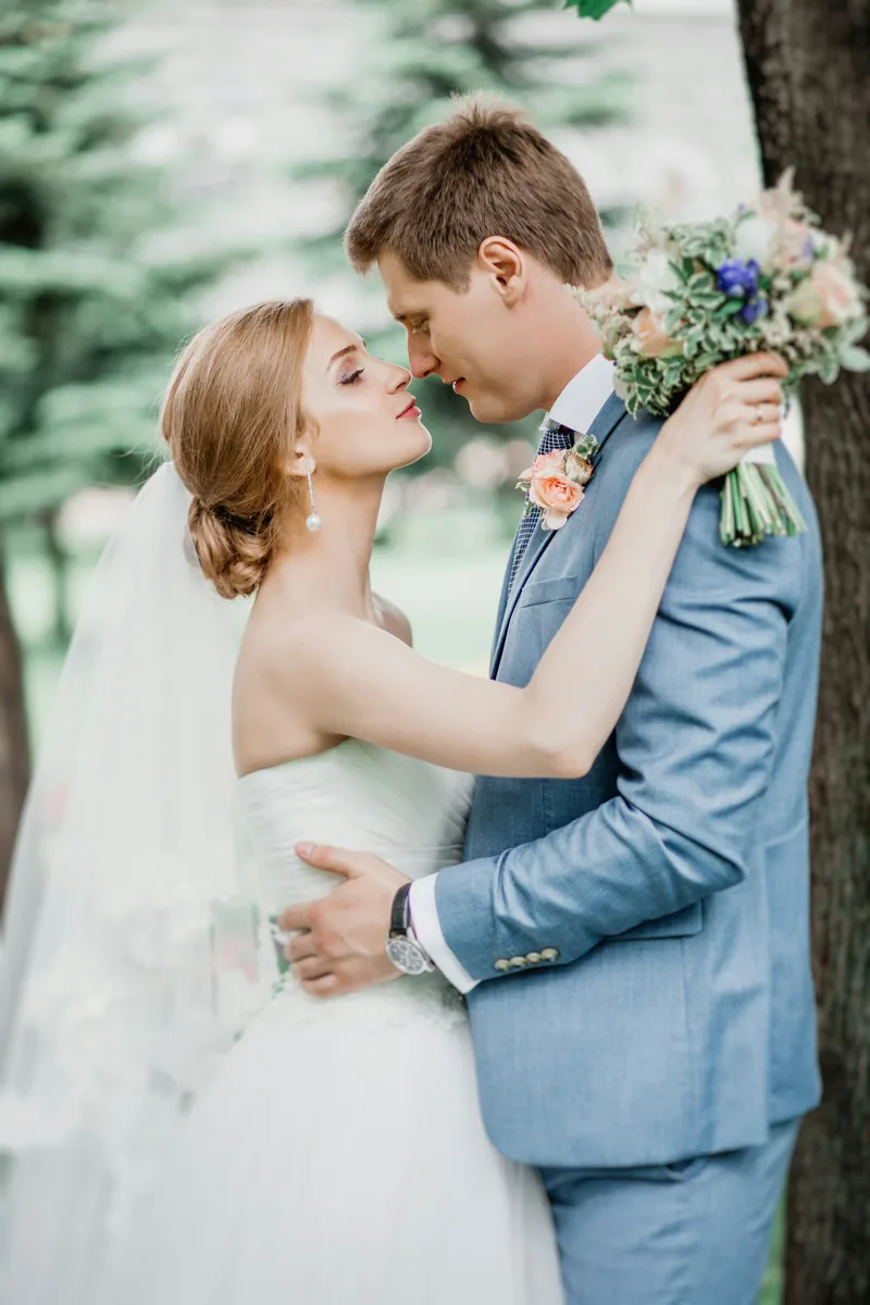 A bride and groom close together among trees, the bride holding a pastel bouquet