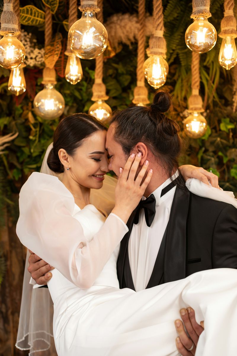 A couple embracing beneath hanging Edison bulbs against a wall of greenery