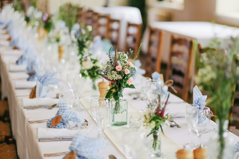 A long table set with white linen, blue gingham napkins and jars of wildflowers