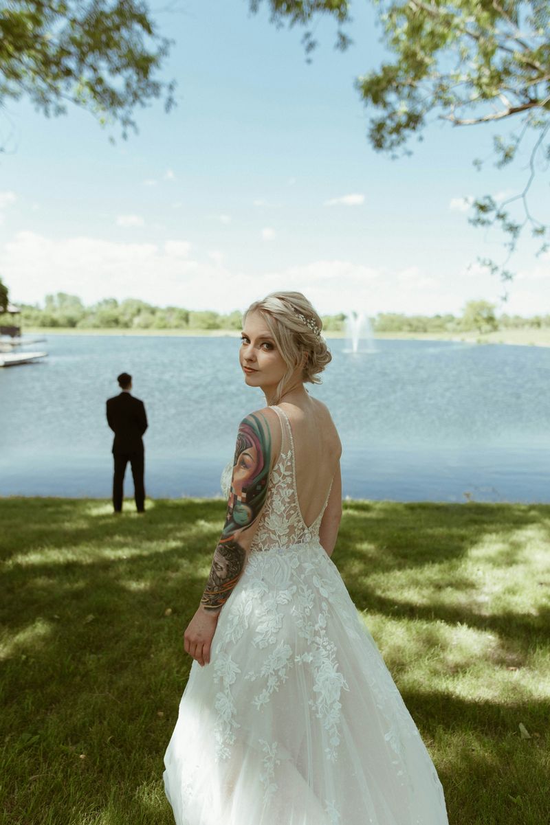 A bride looking back toward the camera on a lawn beside a lake with a fountain
