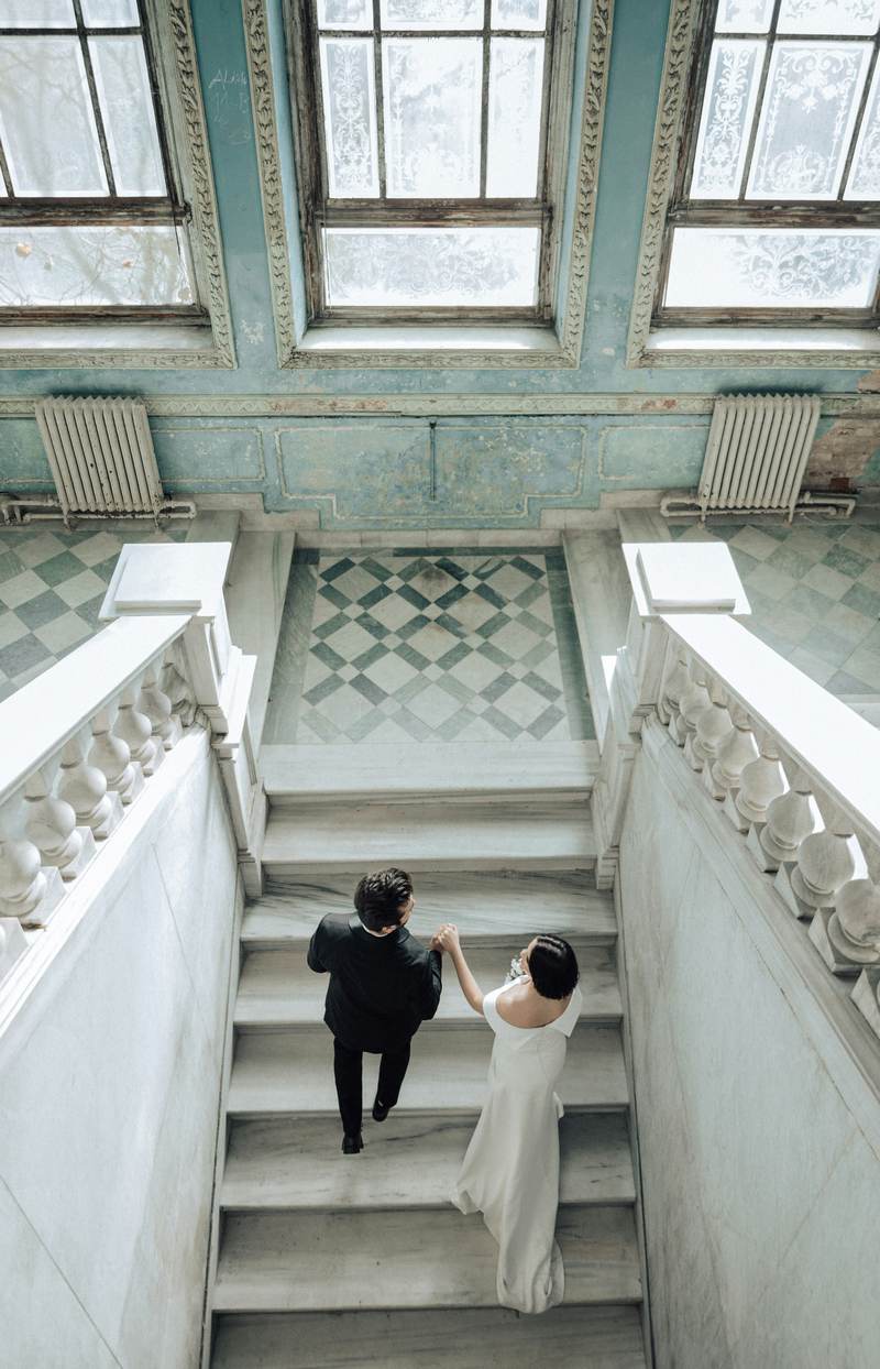 A couple walking up a sweeping marble staircase beneath tall arched windows