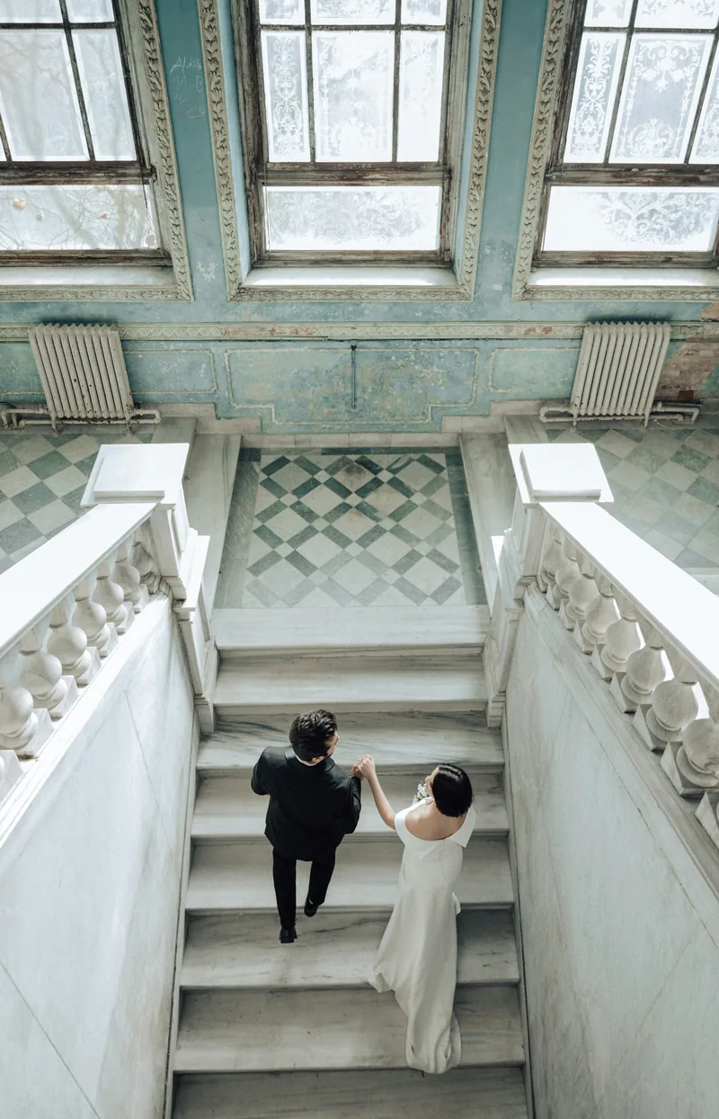 A couple walking up a sweeping marble staircase beneath tall arched windows