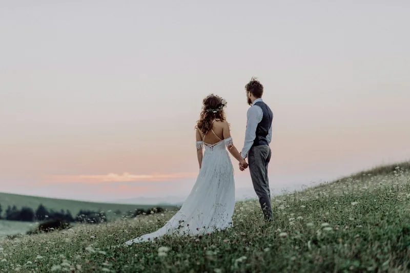 A couple holding hands in a wildflower meadow at dusk beneath a pale pink sky