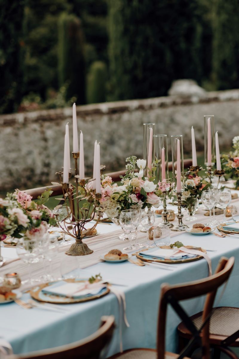 A long table dressed in pale blue linen with pink tapers beside an old stone garden wall