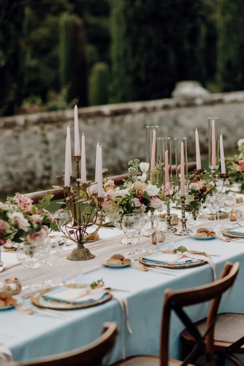 A long table dressed in pale blue linen with pink tapers beside an old stone garden wall