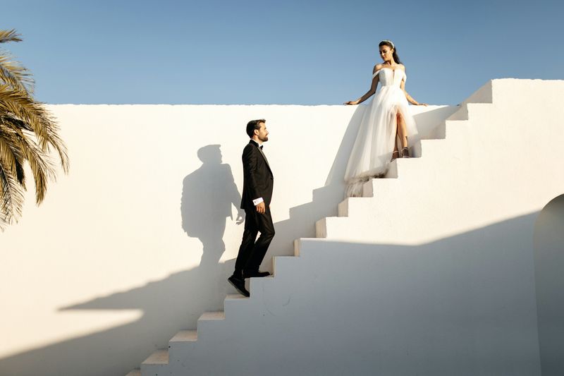 A bride at the top of whitewashed steps as the groom climbs toward her under a bright sky