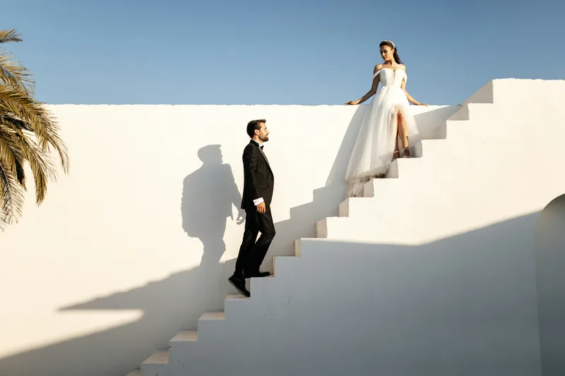 A bride at the top of whitewashed steps as the groom climbs toward her under a bright sky